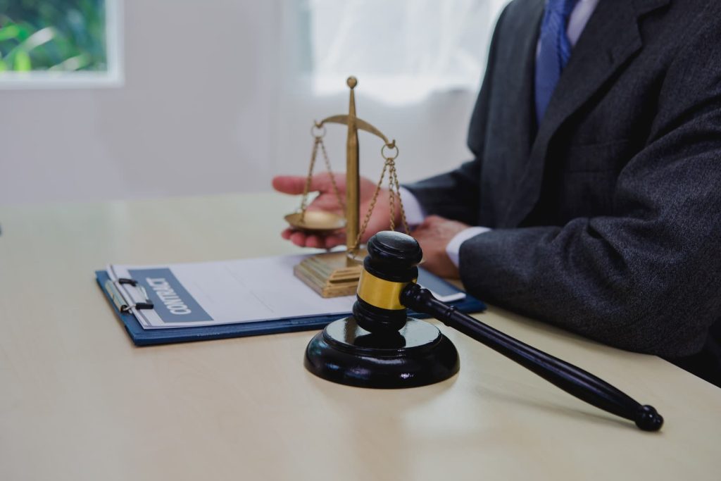 A legal professional in a suit sits at a desk featuring a wooden gavel, a gold scale of justice, and a contract on a clipboard.