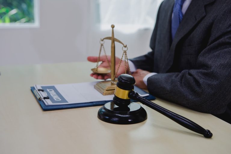 A legal professional in a suit sits at a desk featuring a wooden gavel, a gold scale of justice, and a contract on a clipboard.